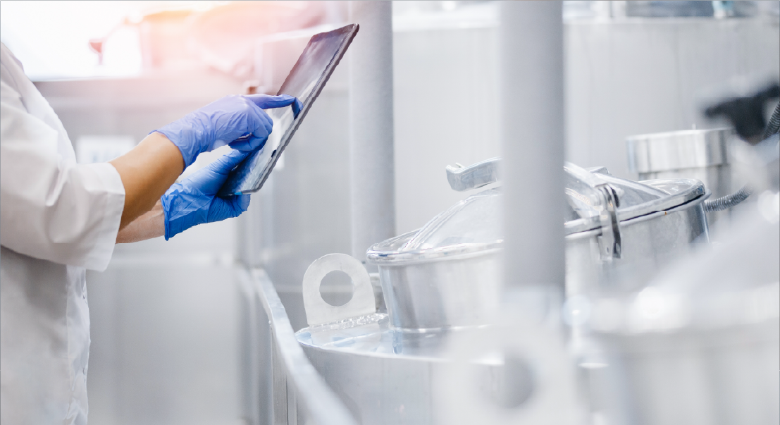 Laboratory technician wearing blue gloves uses a tablet beside stainless steel processing tanks in a clean facility.