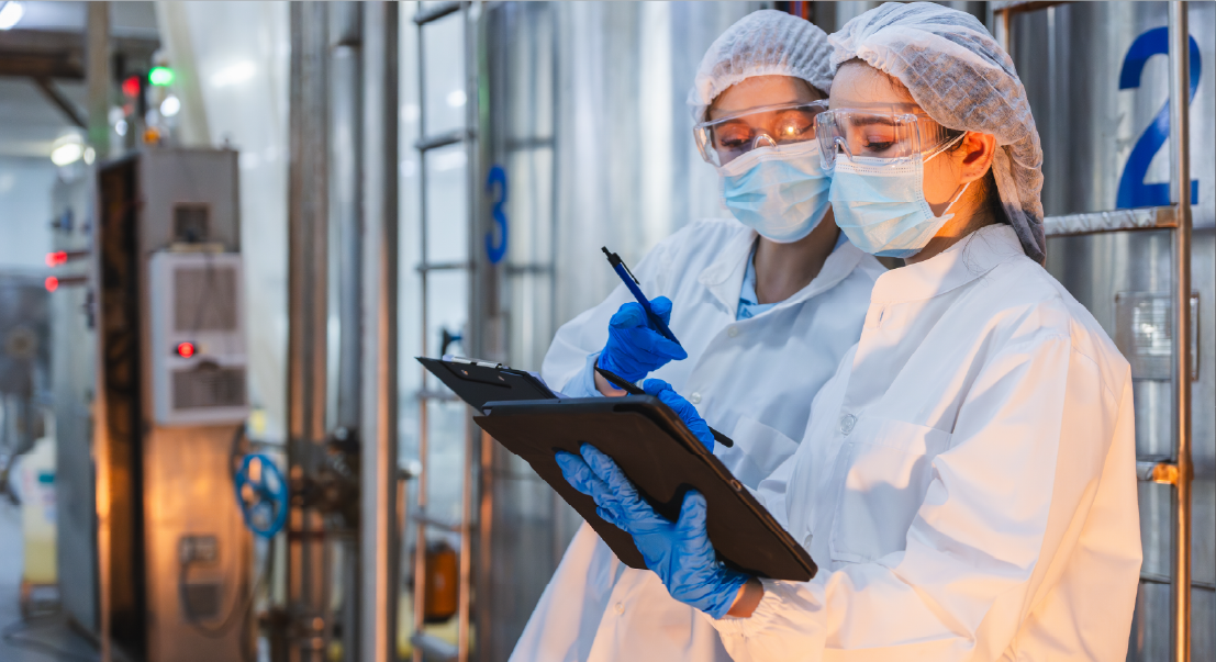 Two technicians in lab coats, gloves, masks, and hairnets review data on a clipboard in a sterile industrial setting.