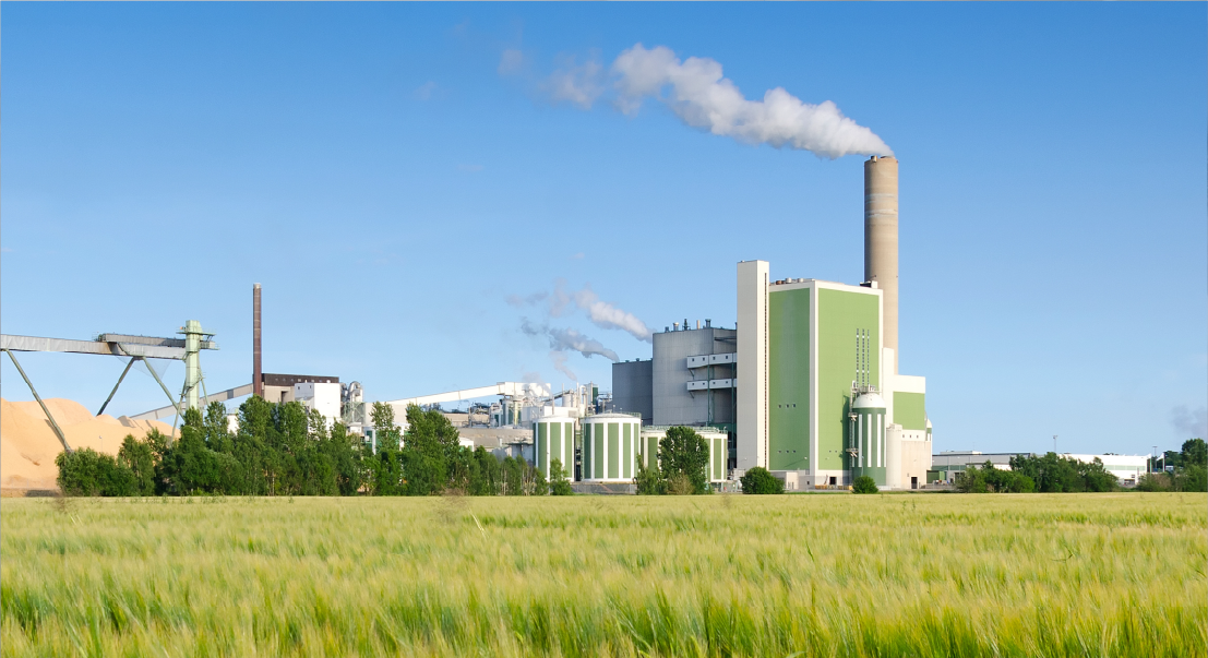 Industrial power plant with a tall smokestack, green and gray buildings, and a line of trees under a blue sky.